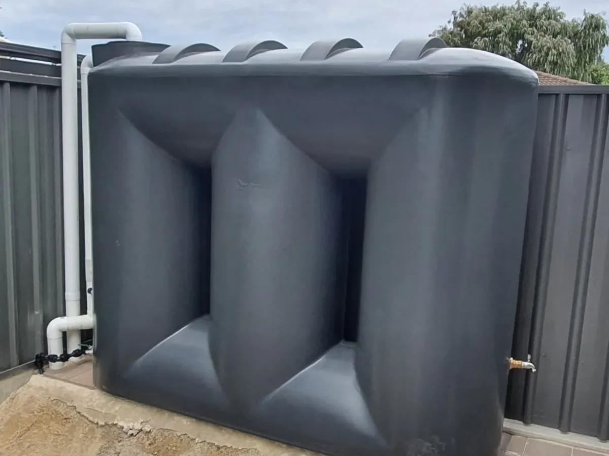 Charcoal slimline rainwater tank against a Colorbond fence, with white PVC inlet pipework on the left and a brass outlet tap at its base