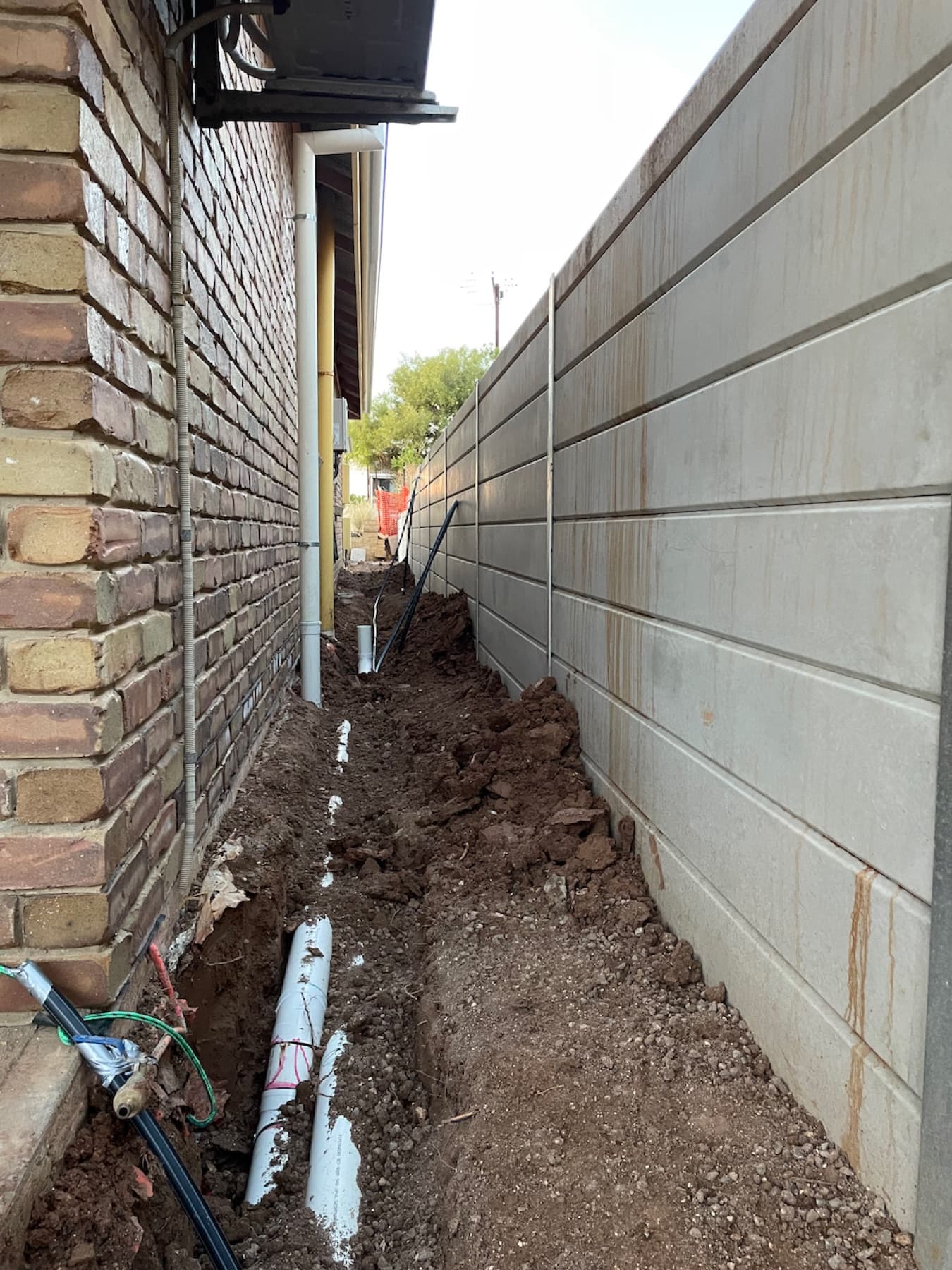 Narrow side passage between house and retaining wall with trench dug for downpipe connection