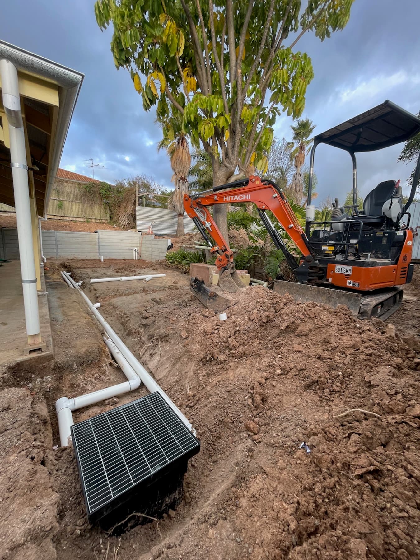 Mini excavator digging near a house with new drainage grate and pipe visible in the excavated ground