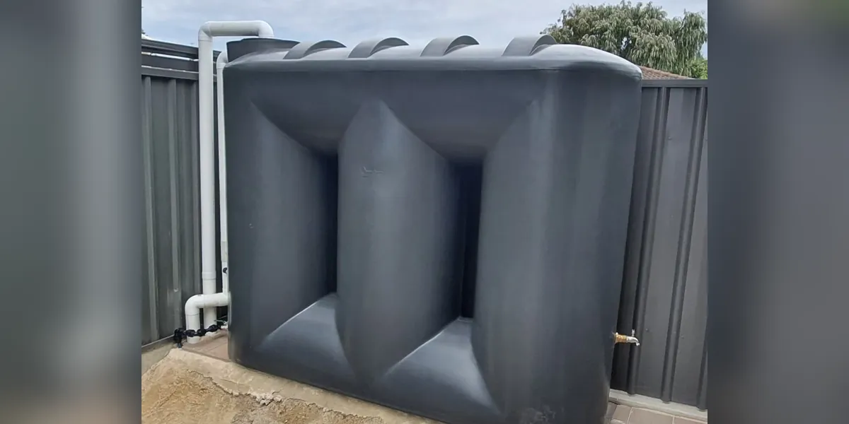 Charcoal slimline rainwater tank against a Colorbond fence, with white PVC inlet pipework on the left and a brass outlet tap at its base