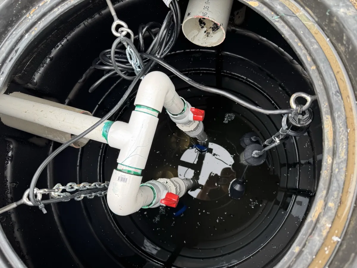 Looking down into a sump chamber at a white PVC pump manifold with isolation valves and float switches